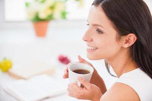 My morning coffee. Side view young smiling woman sitting in bed and holding a cup while covered with blanket photo