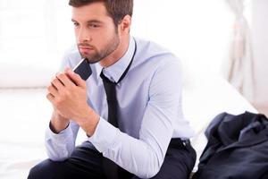 Lost in business thoughts. Thoughtful young man in shirt and tie touching his chin with mobile phone and looking away while sitting on the bed photo