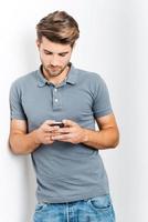 Texting to friend. Handsome young man holding mobile phone and looking at it while leaning at the wall photo