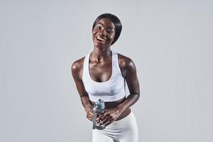 Beautiful young African woman in sports clothing holding bottle with water and smiling photo