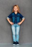 Cheerful girl. Full length of cheerful little girl holding hands on hips and looking at camera with smile while standing against grey background photo