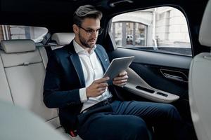 Concentrated at work. Confident young man in full suit working using digital tablet while sitting in the car photo