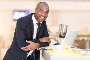 Business on the go. Cheerful young African man in formalwear using his laptop while leaning at bar photo