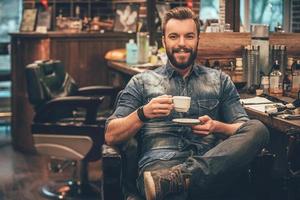 taza de café recién hecho en la barbería. alegre joven barbudo mirando a la cámara y sosteniendo una taza de café mientras se sienta en una silla en la barbería foto