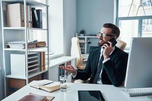 Handsome young man in shirt and tie talking on the smart phone and smiling while sitting in the office photo