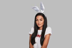 Playful beauty. Attractive young woman in bunny ears looking at camera and smiling while standing against grey background photo