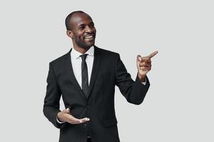 encantador joven africano en ropa formal señalando el espacio de la copia y sonriendo mientras está de pie contra el fondo gris foto