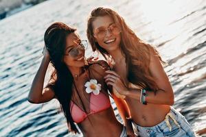 Summer fun. Two attractive young women in swimwear smiling and looking away while standing near the river outdoors photo