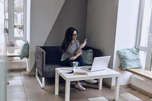 mensaje del cliente. bella joven con anteojos sosteniendo un teléfono inteligente y mirándolo con una sonrisa mientras se sienta en el sofá de su lugar de trabajo foto