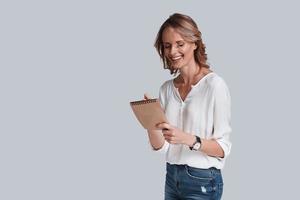 Making notes. Beautiful young woman in casual wear writing something down and smiling while standing against grey background photo