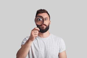 Modern detective. Playful young man applying magnifying glass while standing against grey background photo