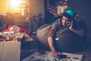 Handsome young man talking on mobile phone and holding joystick while lying on bean bag in messy room after party photo