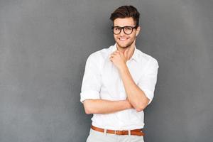 Keep smiling Young handsome man keeping hand on chin and looking at camera with smile while standing against grey background photo