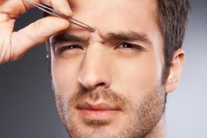 Man tweezing eyebrows. Frustrated young man tweezing his eyebrows and looking at camera while standing isolated on grey background photo