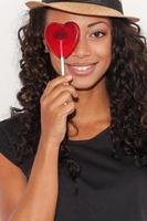 Beauty with candy. Playful young African woman in funky hat holding heart shape candy in front of her eye and smiling while standing against white background photo