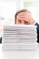 Tired and overworked. Depressed young man in shirt and tie looking out of the stack of documents laying on the table and touching his forehead with hand photo