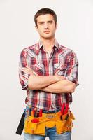Confident handyman. Handsome young handyman with tool belt keeping arms crossed and looking at camera while standing against grey background photo