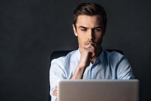 concentrado en el trabajo. un joven apuesto y confiado mirando su laptop y manteniendo la mano en la barbilla mientras se sienta contra el fondo negro foto