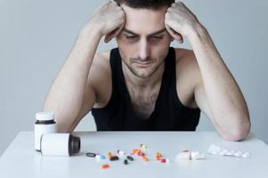 Addicted to these pills. Frustrated young man sitting at the table and looking at the pills laying on it photo