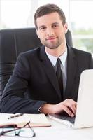 Confident and successful businessman. Handsome young man in formalwear working on laptop and smiling while sitting at his working place photo