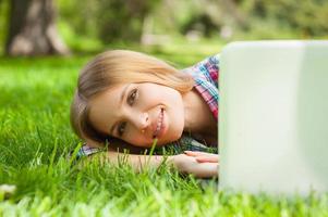 Relaxing in park. Beautiful young female student looking out of laptop monitor and smiling while lying on the grass in park photo