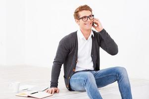 Good talk. Handsome young man talking on the mobile phone while sitting on the floor with note pad and cup of coffee near him photo