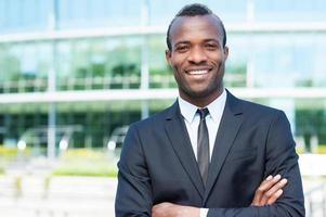 Confidence and success. Confident young African man in full suit keeping arms crossed and looking at camera while standing outdoors photo