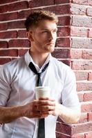 tomando un café. apuesto joven con camisa y corbata sosteniendo una taza de café y mirando por la ventana foto