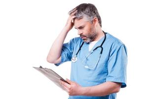 Bad news. Frustrated mature surgeon in blue uniform holding hand in hair and looking at his clipboard while standing isolated on white photo