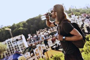 Feeling thirsty. Handsome young man in casual clothing drinking water while standing on the hill outdoors photo