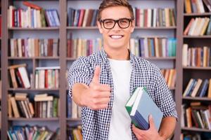 le encanta estudiar. apuesto joven sosteniendo libros y mostrando su pulgar hacia arriba mientras está de pie en la biblioteca foto