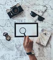 Trying to see everything. Close up top view of man using magnifying glass on copy space with sunglasses, photo camera, compass, passport lying on map around