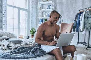 navegar por sitios web favoritos. apuesto joven africano cubierto con toalla de ducha sonriendo y usando la computadora mientras se sienta en la cama en casa foto