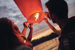 Making happy memories. Happy young people in casual wear holding the sky lantern while standing near the river photo