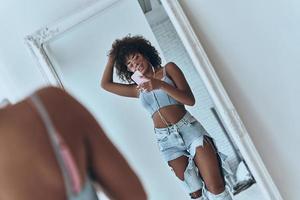 Girl with a great style. Reflection of beautiful young African woman listening music and keeping hand in hair while taking selfie in the mirror at home photo