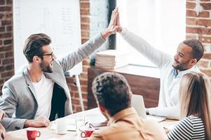 We did it Two young men doing high-five while their coworkers sitting at the office table photo