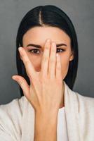 Hide and seek. Portrait of beautiful young woman looking at camera through her fingers while standing against grey background photo