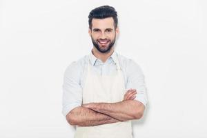 Man in apron. Confident young handsome man in apron keeping arms crossed and smiling while standing against white background photo