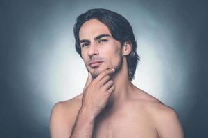 Thoughtful handsome. Portrait of thoughtful young shirtless man looking at camera and holding hand on chin while standing against grey background photo