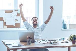 This is success Cheerful young handsome man keeping arms outstretched and keeping eyes closed with smile while sitting at his working place photo