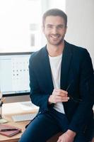 Confident business expert. Confident young man in smart casual wear carrying eyeglasses and smiling while leaning at the office desk photo