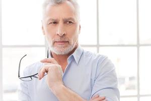 I think I have an idea. Thoughtful grey hair senior man in shirt holding his eyeglasses and looking away while standing in front of the window photo