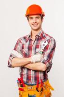 Ready to work. Handsome young handyman with tool belt holding wrench and smiling while standing against grey background photo