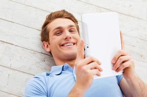 Examining his new gadget. Top view of happy young man lying on the floor and holding digital tablet photo