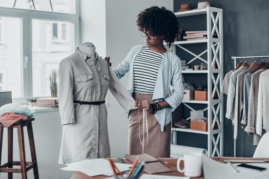 joven perfeccionista. hermosa joven africana midiendo la manga con cinta mientras está de pie en su taller con ropa colgada en el fondo foto