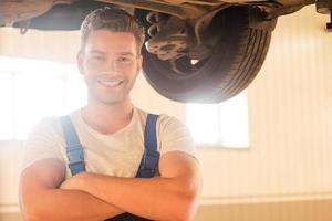 Confident professional. Handsome young man in uniform keeping arms crossed and smiling while standing underneath a car in workshop photo