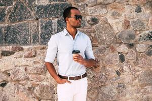 Enjoying fresh coffee outdoors. Handsome young African man in smart casual clothes holding coffee cup while standing against the stoned wall outdoors photo