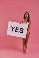 Quick answer.  Full length of attractive young woman holding a poster and smiling while standing on tiptoe against pink background photo