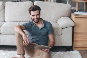 Spending great time at home. Handsome young man holding his digital tablet and looking at camera with smile while sitting on the carpet at home photo