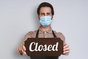 Confident young man in apron wearing protective face mask and holding close sign while standing against gray background photo